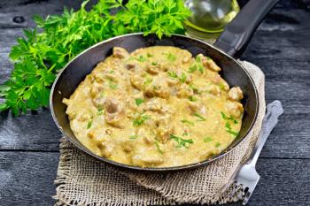 Meat stewed with cream in an old frying pan on burlap, parsley on a black wooden board background