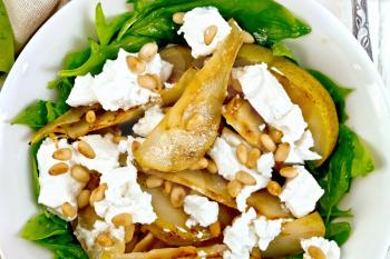 Salad of fried pear, spinach, salted feta cheese and cedar nuts in a plate on a napkin against a light wooden board on top