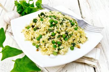 Couscous with spinach and green peas in a plate on a kitchen towel, basil and fork on a light wooden plank background