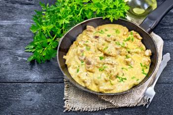 Meat stewed with cream in an old frying pan on burlap, parsley, fork on a black wooden board background
