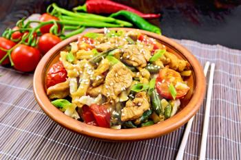 Thai noodles wok with chicken meat, tomatoes, soy sauce and green beans sprinkled with sesame seeds in a plate on a bamboo napkin on a background of a dark wooden board