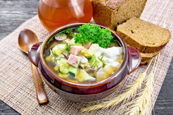 Cold soup okroshka from sausage, potatoes, eggs, radish, cucumber, greens and drink of kvass in a clay bowl, bread on napkin on background of dark wooden board