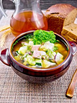 Cold soup okroshka from sausage, potato, egg, radish, cucumber, greens and drink of kvass in a clay bowl, bread on napkin on wooden board background