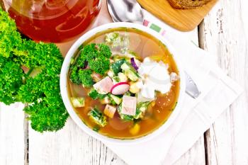 Cold soup okroshka from sausage, potatoes, eggs, radish, cucumber, greens and kvass in a white bowl on napkin, bread and jug with drink on the background of wooden board from above