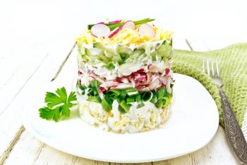 Salad layered from a radish, cucumber, eggs, green onions and cheese in plate, napkin and fork on wooden board background

