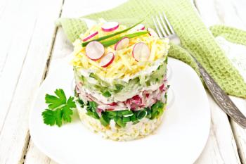 Salad layered from a radish, cucumber, eggs, green onions and cheese in plate, napkin and fork on light wooden board background