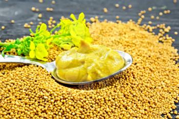 Mustard sauce in a metal spoon with a yellow flower on mustard seeds against a black wooden board