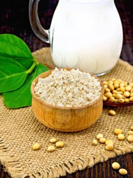 Soy flour in the bowl, soybeans in a spoon and on a napkin of burlap, milk in a jug, green soya leaf against a dark wooden board