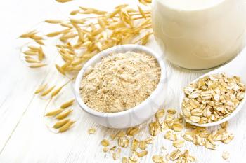 Flour oat in bowl, milk in a jug, oatmeal in spoon, oaten stalks against white wooden table