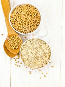 Soy flour and soybeans in two white bowls, spoons on a wooden board background from above