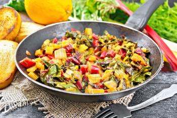 Warm chard salad with orange and onion in a frying pan on sackcloth, bread, fork on a dark wooden board background
