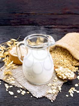 Oat milk in jug, flour in bowl, oatmeal in spoon, grain in jute bag, oaten stalks on napkin of burlap on wooden board background