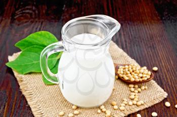Soy milk in a jug, green leaf, soybeans in a spoon and on sackcloth against a dark wooden board