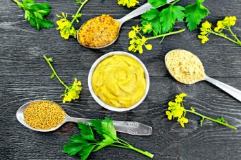 Mustard granular, powder, seeds in spoons and mustard sauce in a bowl, yellow flowers and parsley on the background of a black wooden board from above