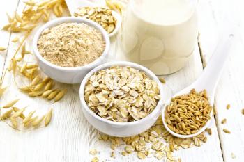 Oat flakes and flour in bowls, grain in a spoon, oatmeal milk in a glass jug and ripe oaten stalks against a white wooden board