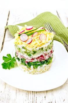 Salad layered from a radish, cucumber, eggs, green onions and cheese in plate, napkin on light wooden board background
