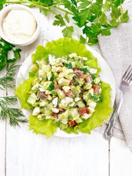 Salad from salmon, cucumber, eggs and avocado with mayonnaise on lettuce leaves in a plate, napkin, dill, parsley and fork on a light wooden background from above