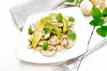 Salad of avocado and raw champignons, seasoned with lemon juice and vegetable oil with mint leaves, kitchen towel and a fork on the background of light wooden board
