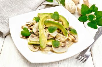 Salad from avocado and raw champignons, seasoned with lemon juice and vegetable oil with mint leaves, napkin and fork on the background of a light wooden board