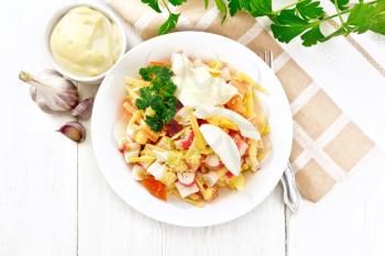 Salad of crab sticks, cheese, garlic, tomatoes and eggs with mayonnaise in a plate, napkin, parsley and fork on a wooden board background from above