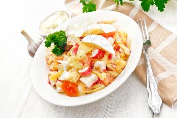 Salad of crab sticks, cheese, garlic, tomatoes and eggs with mayonnaise in a plate, kitchen towel, parsley and fork on a wooden board background
