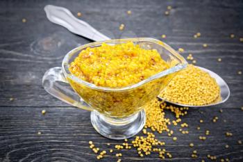 Mustard Dijon sauce in a glass sauceboat and mustard seeds in a spoon on a background of a dark wooden board