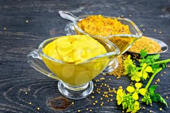 Mustard sauce and Dijon mustard in two glass saucepans, yellow flower and seeds in a spoon on black wooden board background