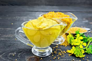 Mustard sauce and Dijon mustard in two glass saucepans, yellow flower and seeds in a spoon on the background of dark wooden board
