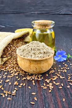 Flax flour in a bowl, seeds in a bag and on a table, a blue linen flower and oil in a glass jar on a background of a dark wooden board