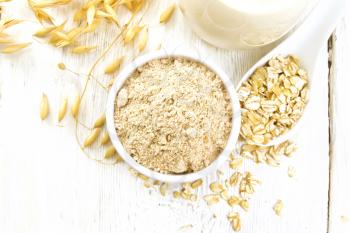 Flour oat in bowl, milk in a jug, oatmeal in spoon, oaten stalks on the background of white wooden board from above

