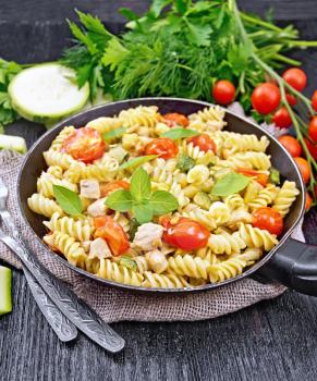 Fusilli with chicken, zucchini and tomatoes in a frying pan on burlap, forks, basil and parsley on a wooden board background