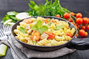 Fusilli with chicken, zucchini and tomatoes in a frying pan on burlap, forks, basil and parsley on dark wooden board background