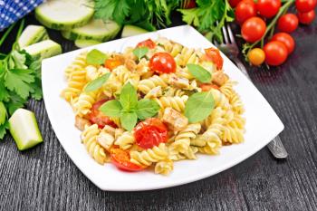 Fusilli with chicken, zucchini and tomatoes in a white plate, napkin, fork, basil and parsley on black wooden board background