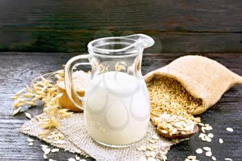 Oat milk in jug, flour in bowl, oatmeal in spoon, grain in jute bag, oaten stalks on burlap against wooden board