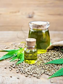 Hemp oil in two glass jars with grain in the sack, leaves and stalks of cannabis on the background of wooden boards