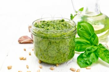Pesto sauce in a glass jar, basil, pine nuts, garlic and olive oil in a carafe on the background of a light wooden board
