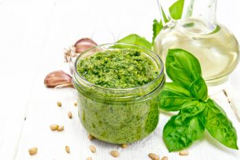 Pesto sauce in a glass jar, basil, pine nuts, garlic and olive oil in a carafe on a wooden board background