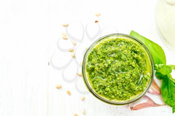 Pesto sauce in a glass jar, basil, pine nuts, garlic and olive oil in a carafe on wooden board background from above