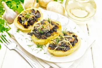 Potatoes stuffed with mushrooms, fried onions and cheese in a plate on towel, vegetable oil in carafe, parsley, garlic and a fork on wooden board background