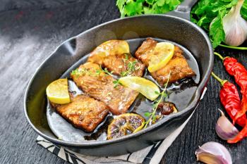 Pink salmon with a sauce of honey, lemon juice, garlic, hot pepper and soy sauce, lemon slices and sprig of thyme in a frying pan on a towel, parsley on black wooden background
