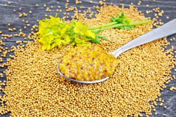 Mustard Dijon sauce in a metal spoon and yellow mustard flower on seeds against a black wooden board