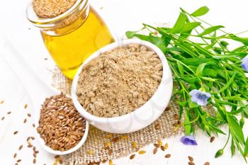 Flaxseed flour in a bowl, linenseeds in a spoon, oil in glass jar on a burlap napkin, leaves and flowers of flax on wooden board background