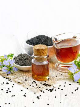 Nigella sativa oil in vial and gravy boat, seeds in a spoon and black cumin flour in a bowl on burlap, kalingi twigs with blue flowers and leaves on wooden board background