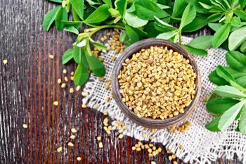 Fenugreek seeds in a bowl on burlap with green leaves on a wooden board background from above