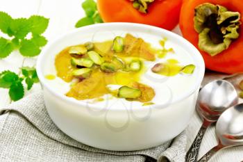 Yogurt, persimmon and honey dessert with vanilla, cardamom and pistachios in a bowl on a napkin, mint and spoon on wooden board background