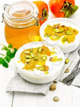 Dessert of yogurt, persimmon and honey with vanilla, cardamom and pistachios in two bowls on a napkin, mint and spoon on background of light wooden board