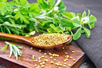Fenugreek seeds in a spoon on a brown plate with herbs, dark napkin on wooden board background