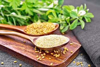 Fenugreek seeds and ground spice in two spoons on a brown plate with herbs, a napkin on black wooden board background