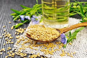 White linen seeds in a spoon, flax stalks with blue flowers and green leaves on burlap, oil in a bottle on wooden board background