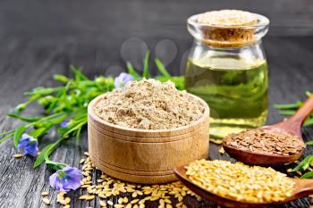 Flaxseed flour in a bowl, white and brown seeds in spoons and on table, flax leaves and flowers, oil in a glass jar on dark wooden board background